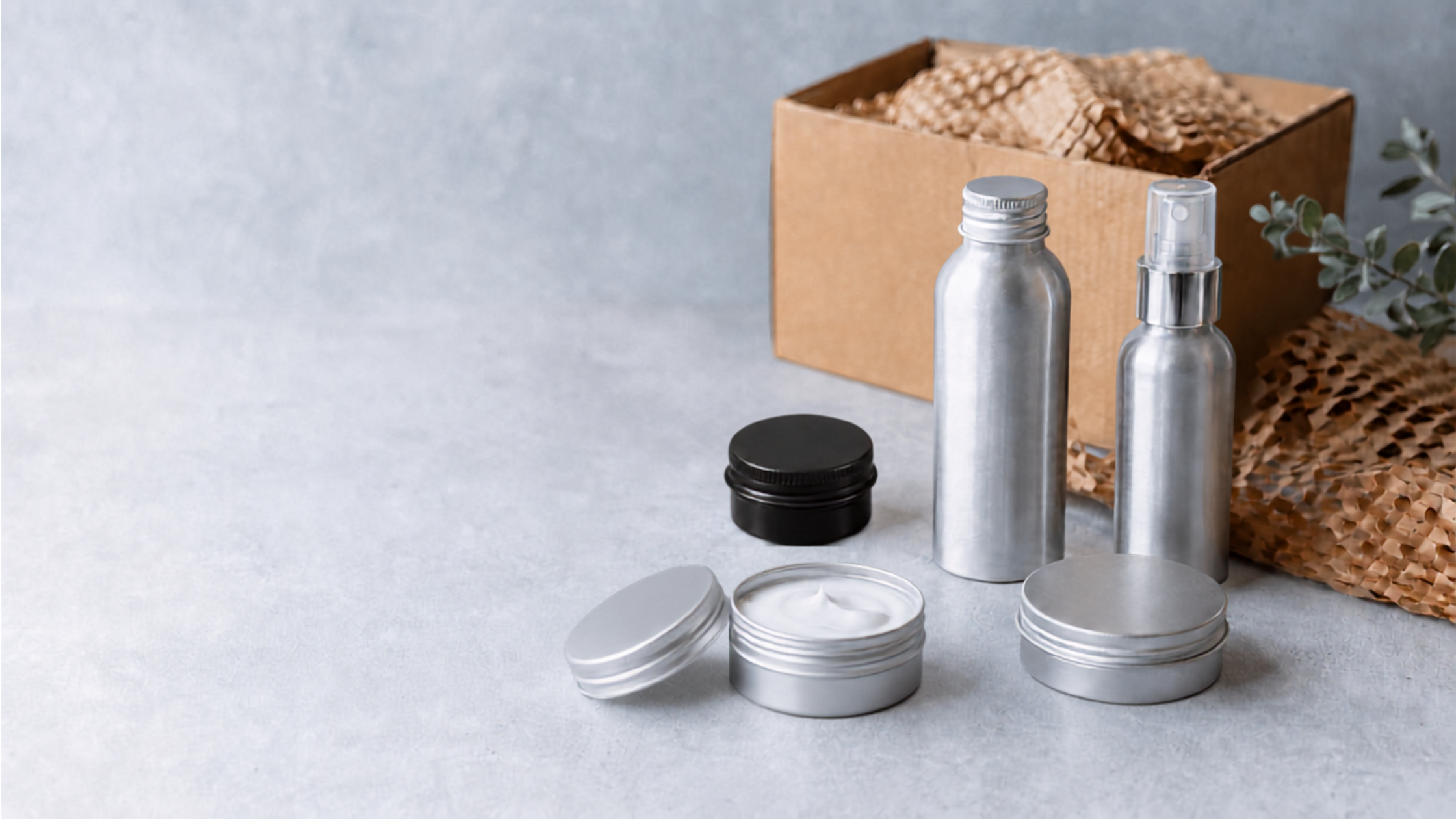 Silver aluminium cosmetic bottles and tins with moisturising cream, presented beside protective paper packaging and a cardboard shipping box.