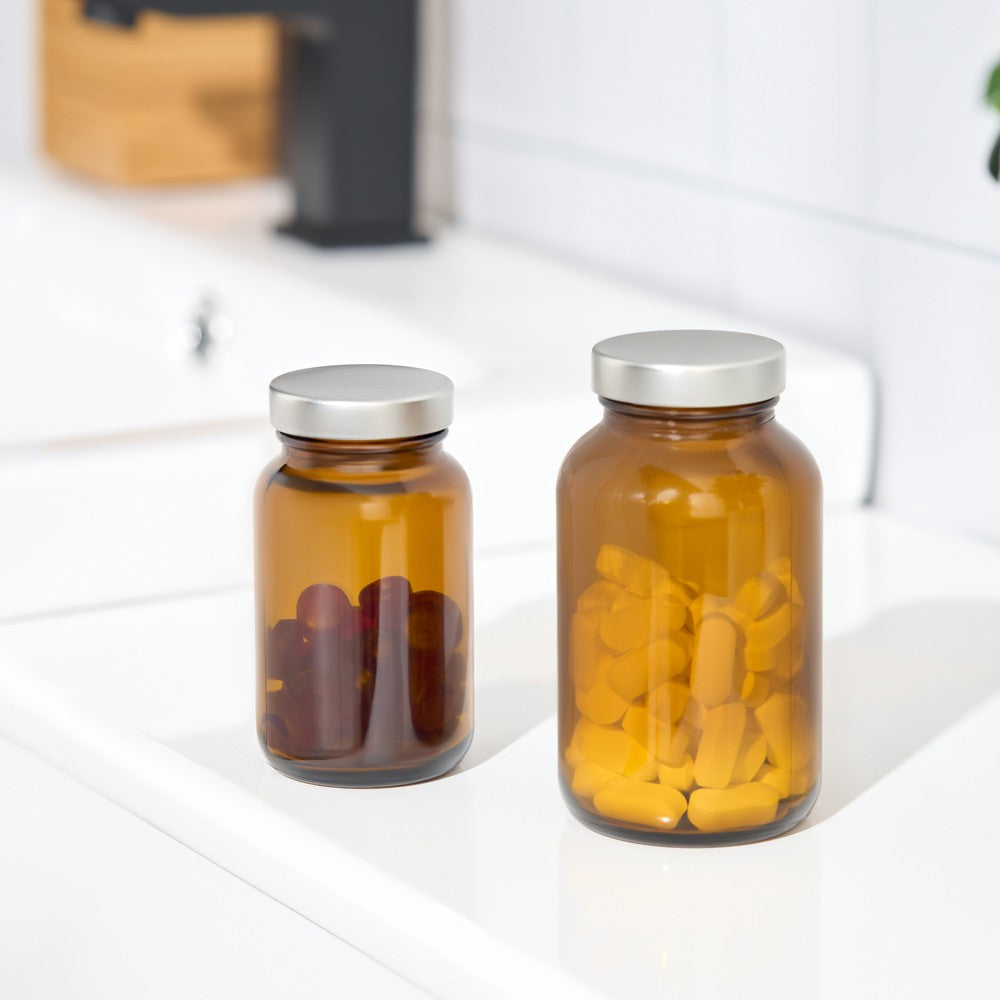 Two amber glass supplement bottles with silver metal lids, filled with capsules, placed on a white countertop in a kitchen setting.