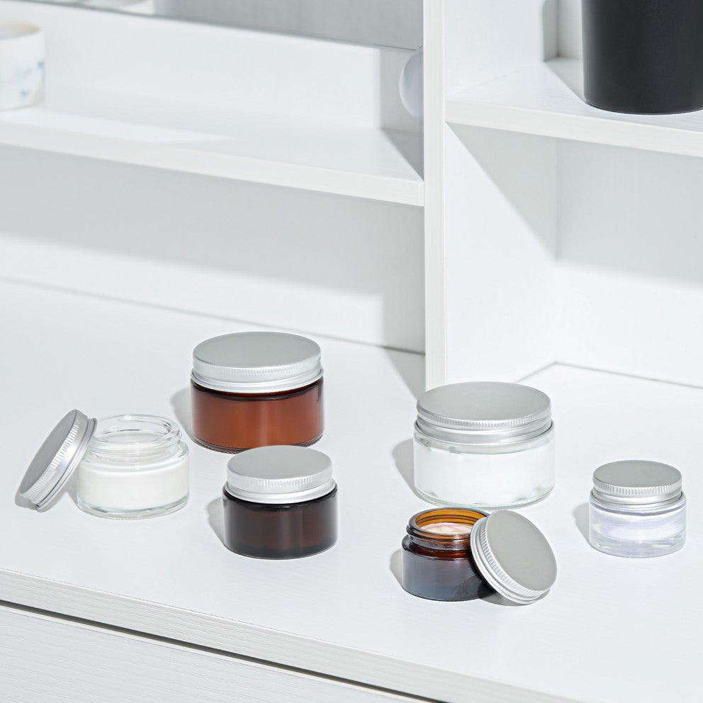 A set of empty cosmetic jars with silver metal lids arranged on a white shelf. The collection includes various sizes of jars in amber brown, clear glass, and white plastic, some open and some closed.