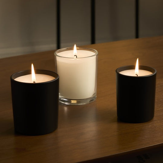 Three lit candles displayed on a wooden table, including two matte black jar candles and one white candle in a clear glass container.