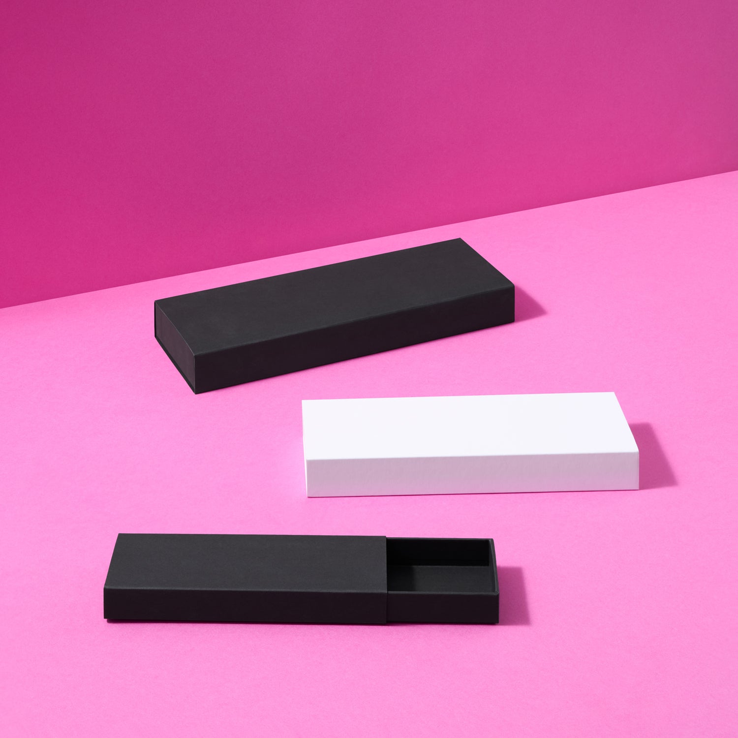 Three rectangular presentation boxes on a bright pink background: one black closed box, one white closed box, and one black open box showing the interior.