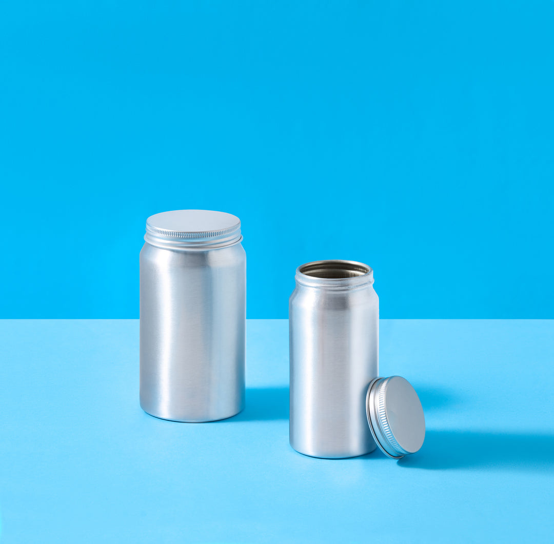 Two empty metallic silver aluminum pill bottles with screw-on caps, one standing upright and the other lying on its side with the cap removed, set against a bright blue and light blue studio background.