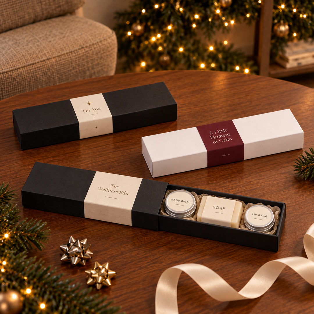 Three gift boxes of bath and body products arranged on a wooden table with holiday decorations in the background. One black box labeled 'The Wellness Edit' is open to show three round soap and balm jars, one white box labeled 'A Little Moments of Calm' is closed, and another black box labeled 'for you' sits behind.