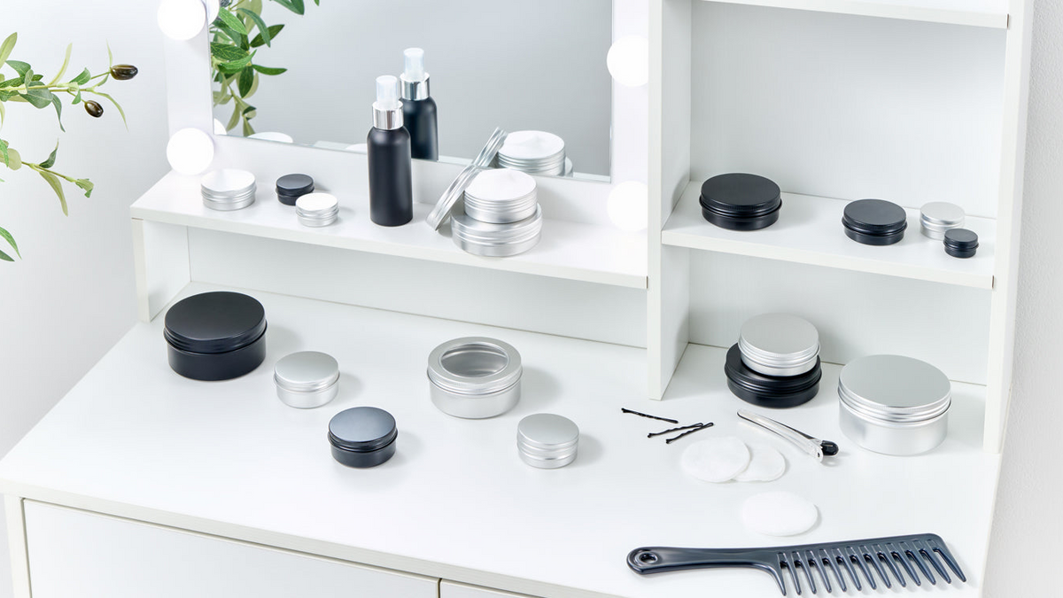 Assorted aluminium and black cosmetic tins displayed on a white vanity table with skincare accessories and a mirror in the background.