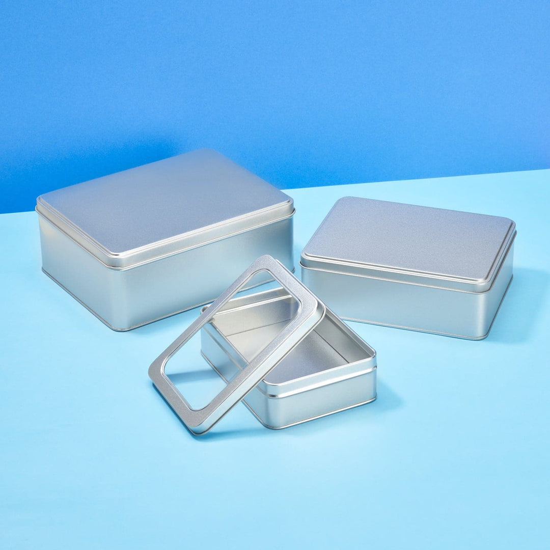 A collection of large silver rectangular tins two with traditional lids and one with a window lid to show the contents inside.