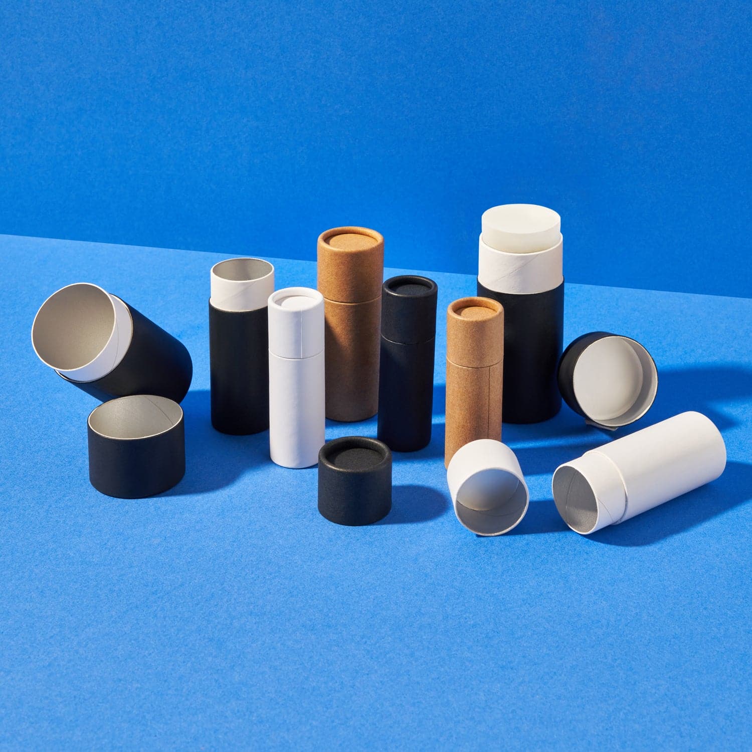 A collection of cardboard tubes with push up bases and internal lining in black, white and brown. Some tubes have lids off and one cardboard tube contains deodorant.