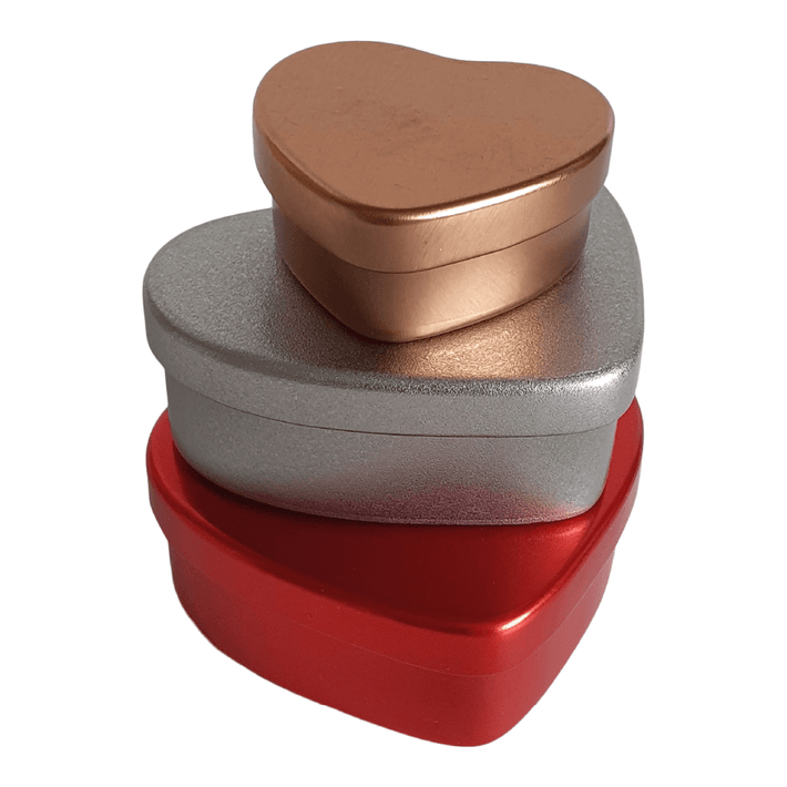 A rose gold heart tin stacked on top of a silver heart shaped tin, which in turn is stacked on top of a red heart tin.